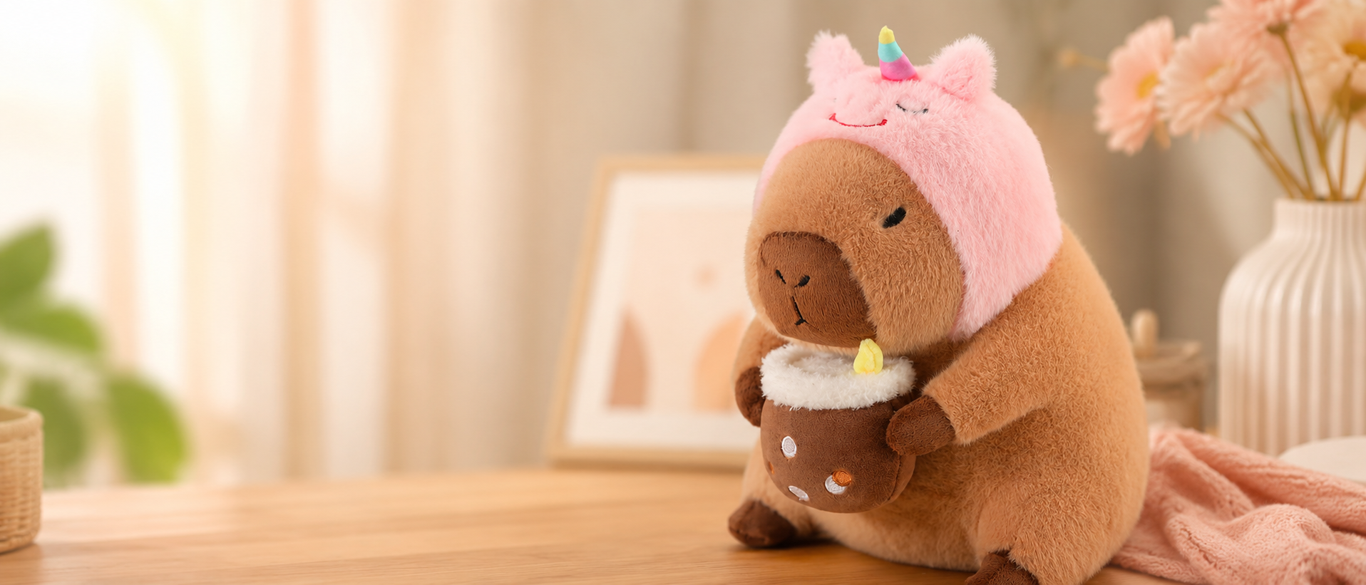 A cute capybara plush toy wearing a pink unicorn hat and holding a small plush drink cup, displayed on a wooden table in a warm cozy room with soft lighting, flowers, and pastel home decor in the background.